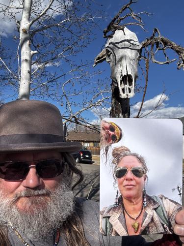 A man with a long gray beard in a hat and sunglasses holds a skull picture in his hands.